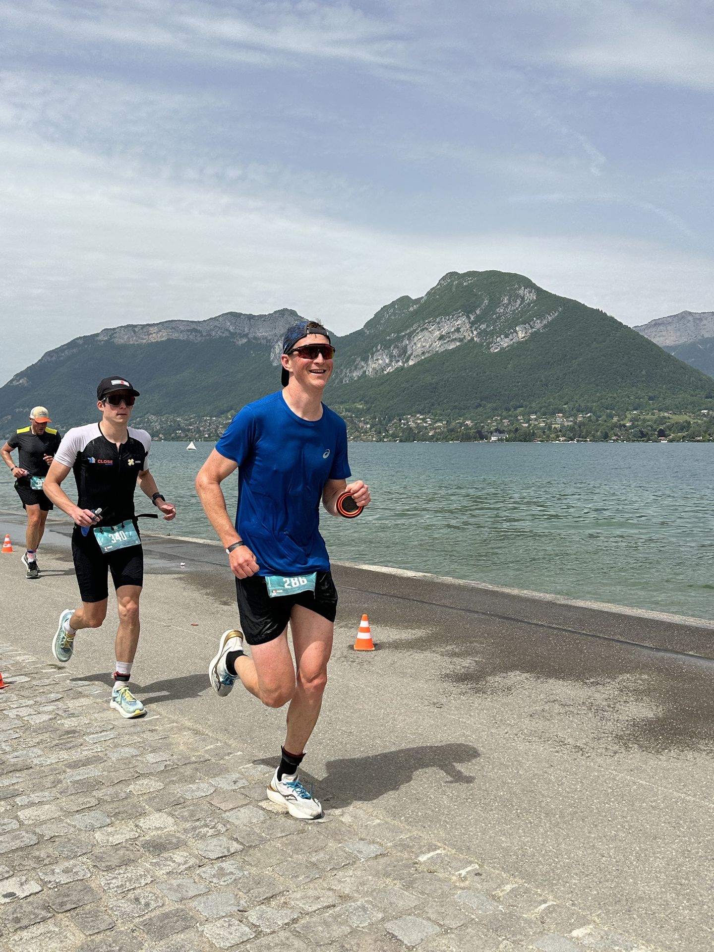 Courir avec le bon équipement pour affronter toutes les conditions Pierre en pleine course sur l'Alpsman, affichant un grand sourire au bord du lac. Équipé d’un t-shirt bleu, d’une casquette et de lunettes, il illustre l’importance d’un équipement adapté pour les longues distances.