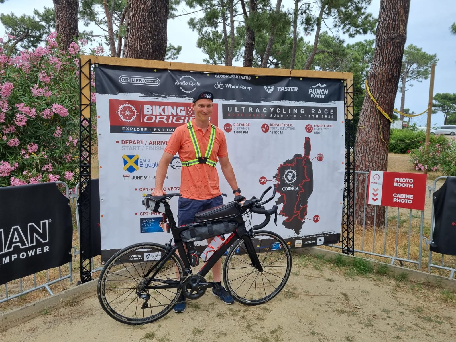 Prêt à affronter 958 km et 16 000 m de D+ en autonomie Pierre prêt pour l’aventure du BikingMan, posant avec son vélo devant l’affiche officielle de la course. Son équipement est optimisé pour l’ultra-distance avec prolongateurs, sacoches et gilet de sécurité.