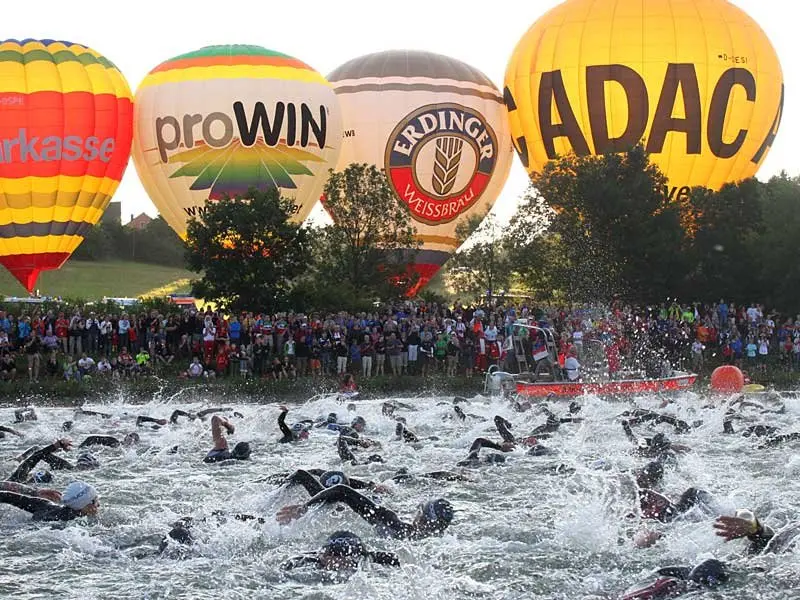 Une bataille aquatique épique sur le point de commencer. Départ de la natation : 3800 m à affronter, une horde de nageurs prêts à plonger, sous le regard du public et des montgolfières en arrière-plan.